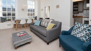 A modern living room with a grey sofa, ottoman, and blue armchair, a dining area with a table and chairs, and a kitchen with a modern stove and cabinets.