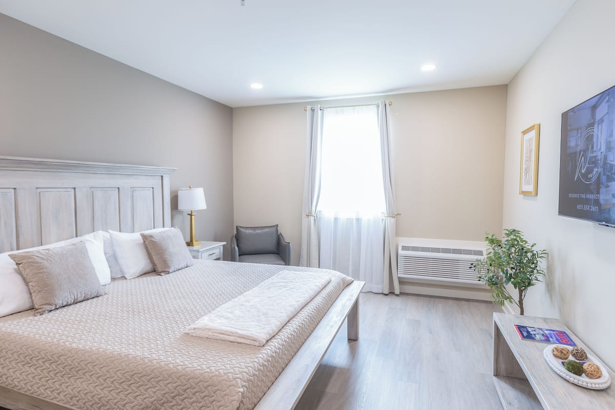 A hotel room with a bed, a bedside table with a lamp, a chair, a window with curtains, an air conditioning unit, and a small table with a plant and a menu.