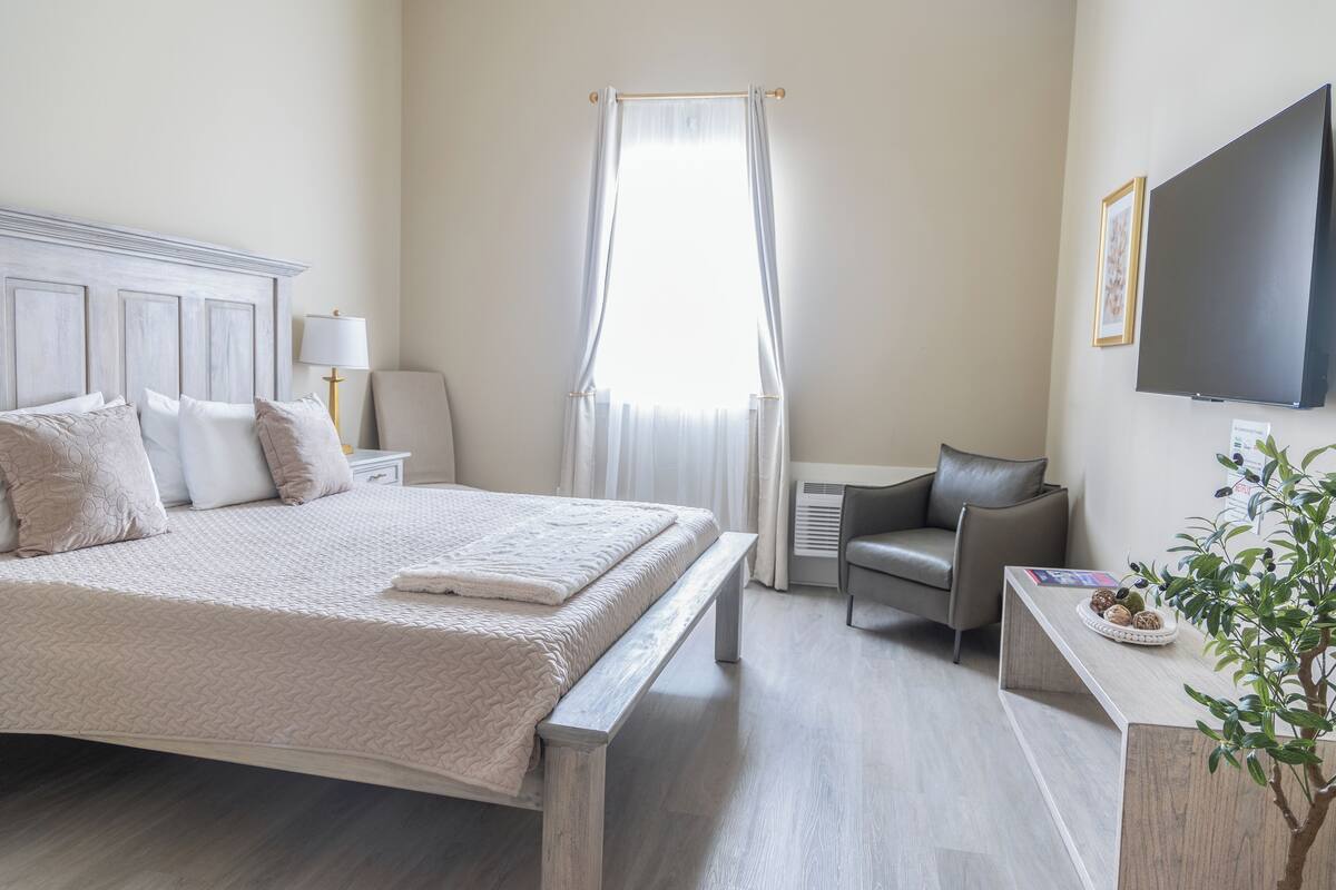 A hotel room with a large bed, a bedside table with a lamp, a chair, a TV mounted on the wall, and a window with curtains.