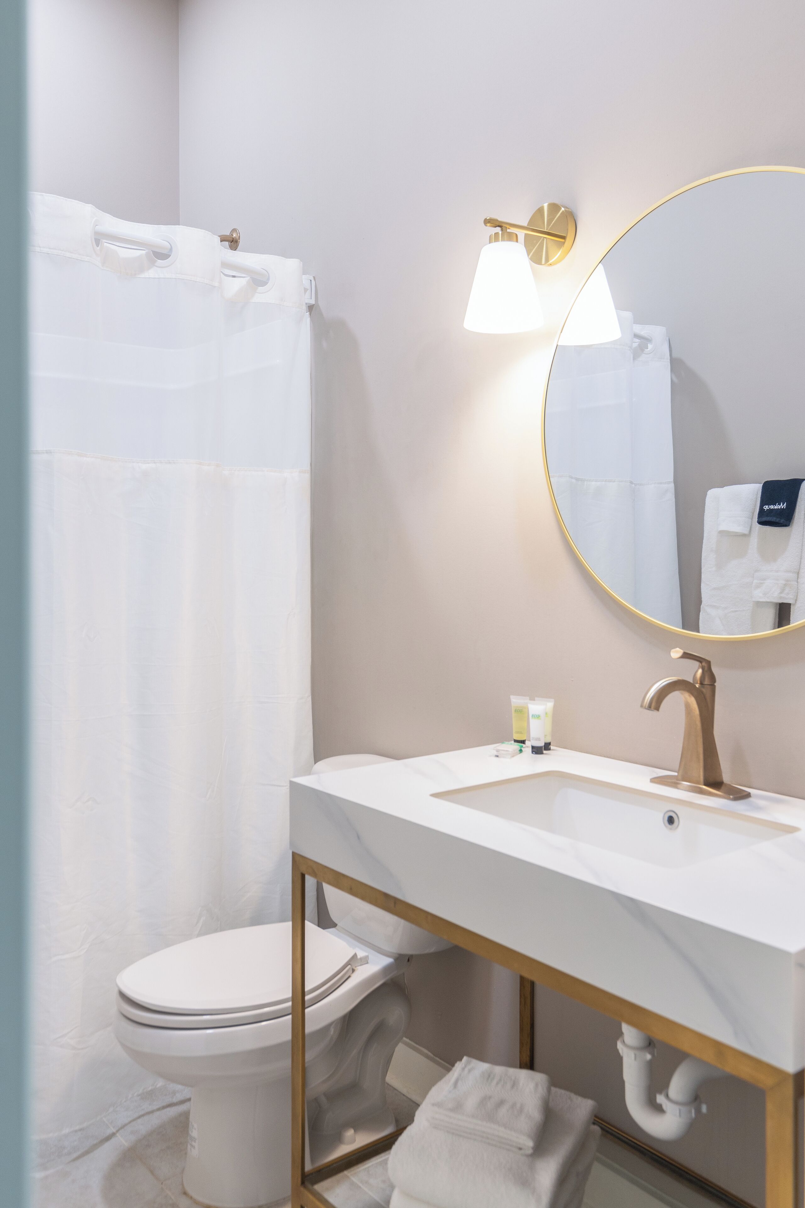A modern bathroom with a white sink, gold fixtures, a round mirror, and a white shower curtain.