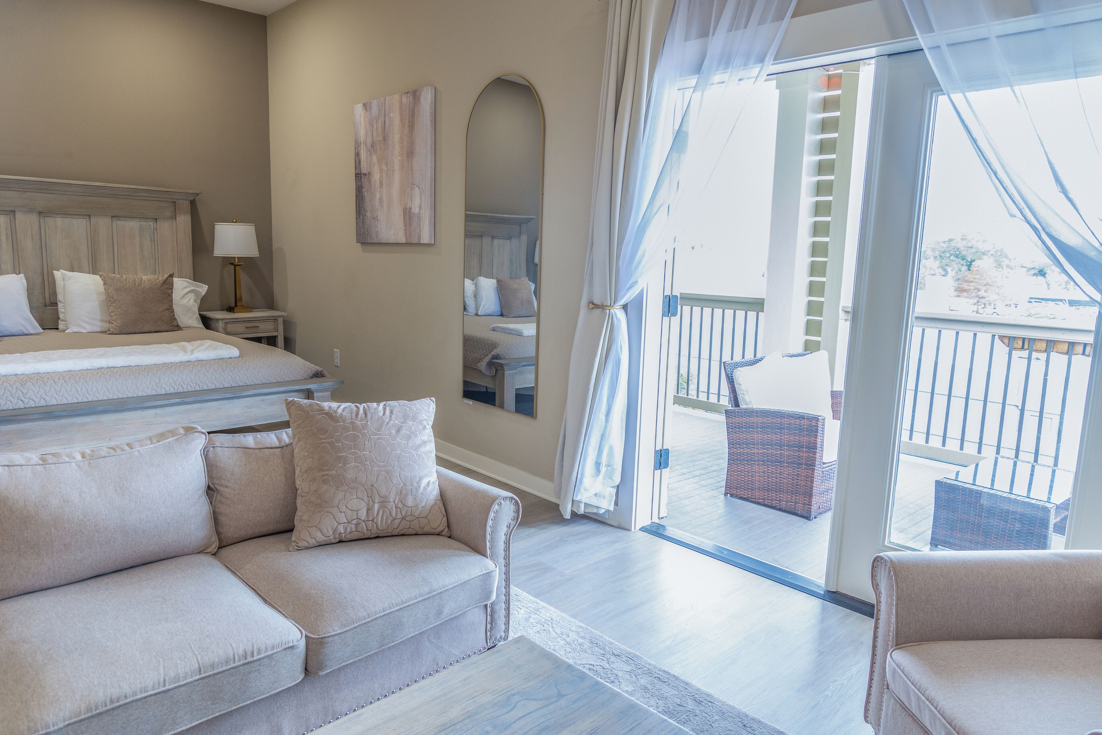 Quarto de hotel com cama, sofá, mesa de cabeceira, luminária, espelho e varanda com mobiliário de vime.
