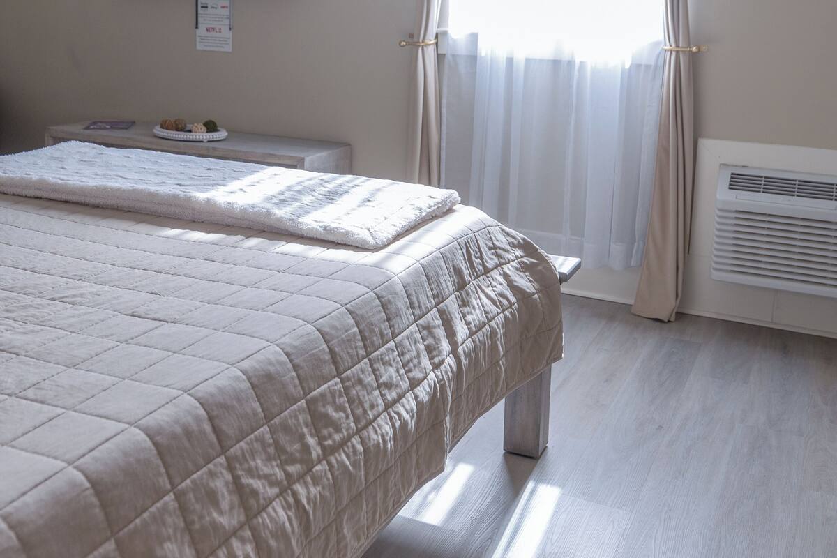 A bedroom with a bed, a flat-screen TV mounted on the wall, a window with sheer curtains, and a small table with a plate and a book.