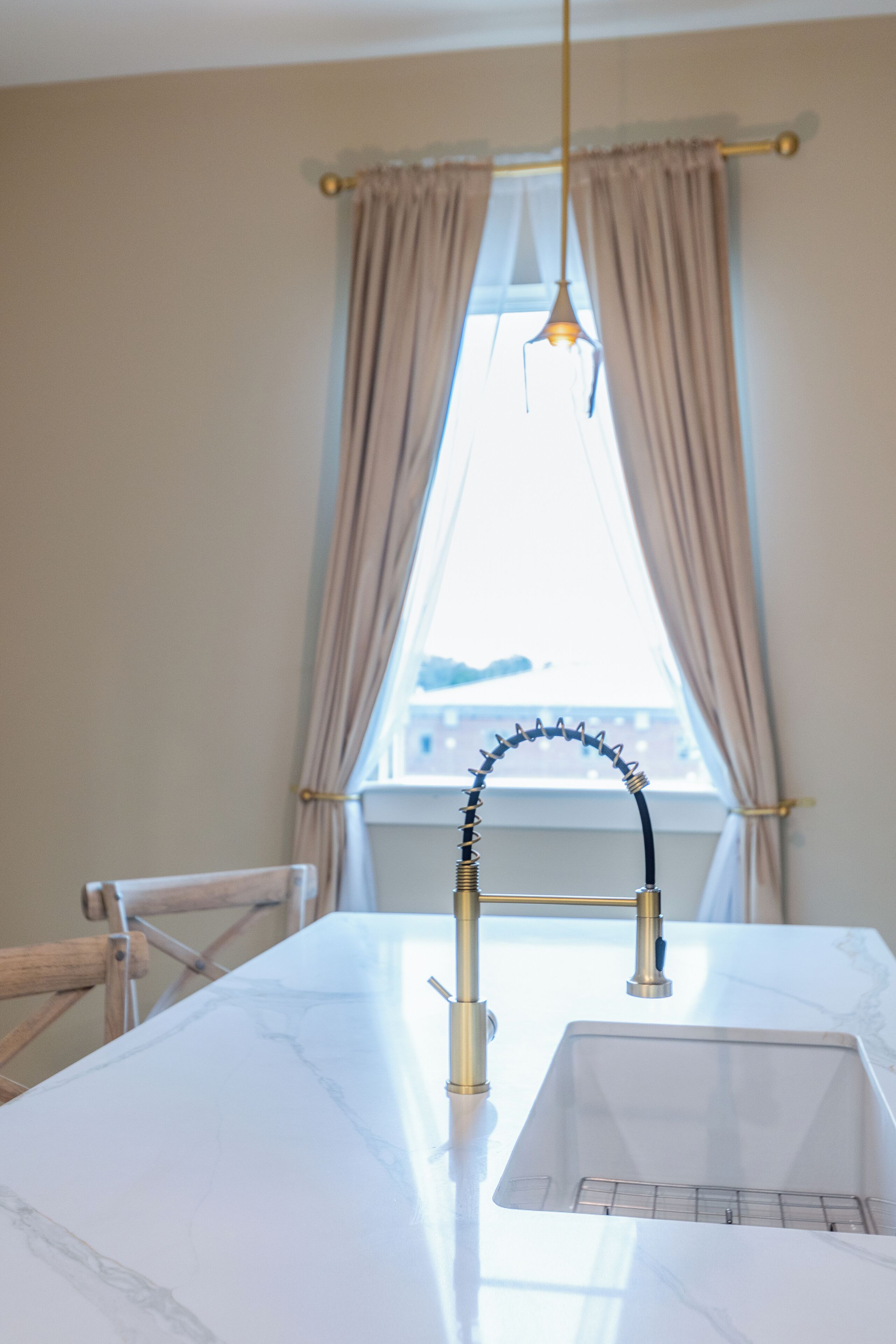Eine Küche mit weißem Marmor-Arbeitsplatz, einem goldenen Wasserhahn und einem Fenster mit beigen Vorhängen.