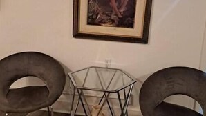 A room with a framed painting on the wall, a hexagonal glass table with a vase, and two dark chairs.