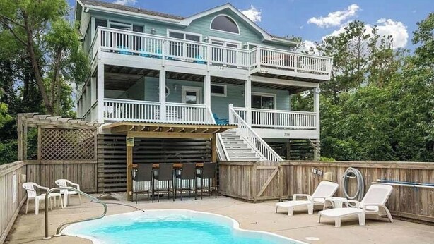 Una casa de dos pisos con piscina, mobiliario exterior y una terraza de madera.