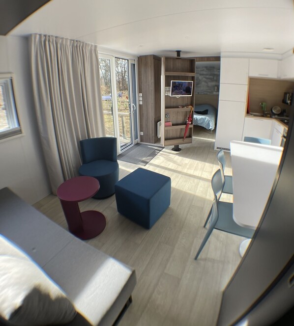 A modern, compact living space with a kitchenette, a seating area with a sofa and ottoman, and a bedroom with a bed and a TV.