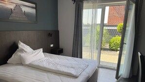 Un lit bien fait, avec du linge de lit blanc, dans une chambre moderne dotée d’une grande fenêtre donnant sur un bâtiment en briques.