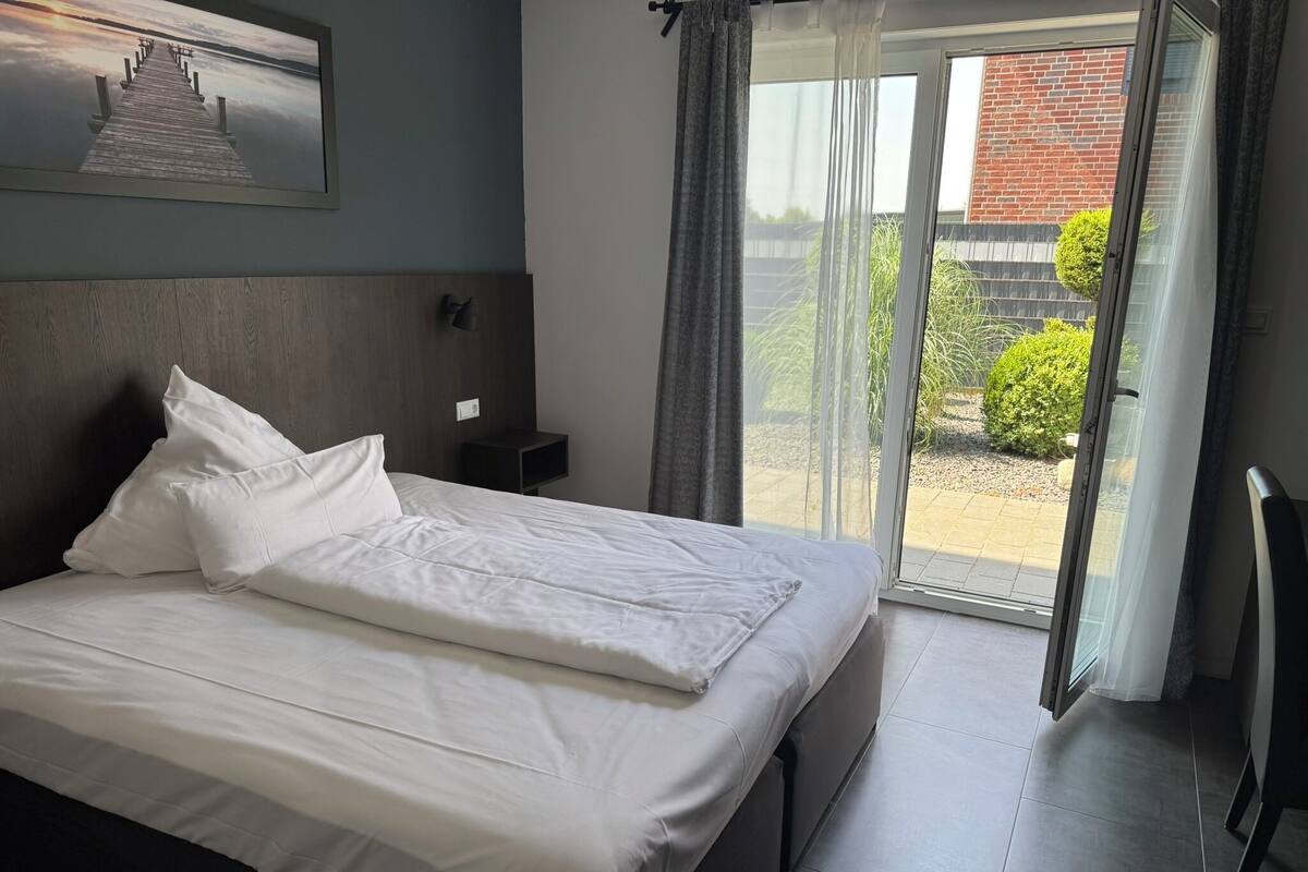A neatly made bed with white linens in a modern room with a large window and a view of a brick building outside.
