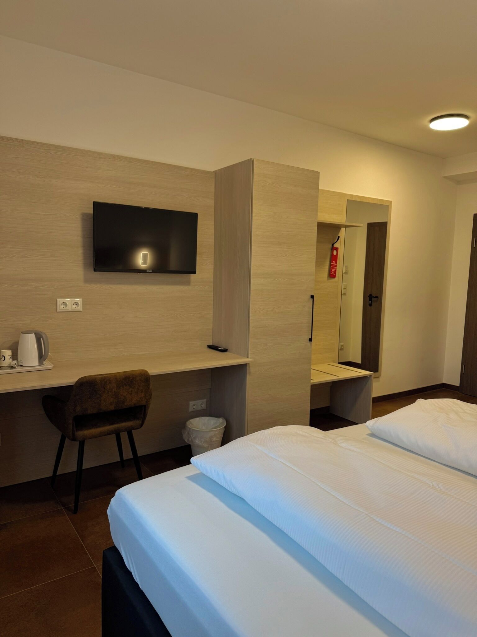 A hotel room with a bed, a desk, a chair, a TV mounted on the wall, and a mirror.