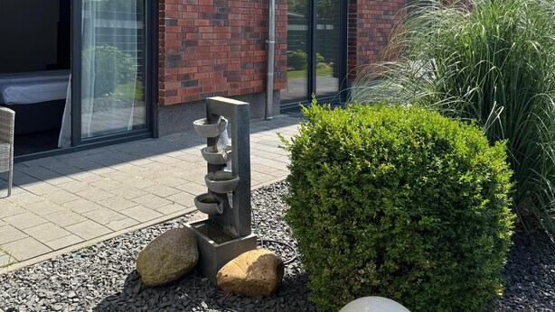 Un bâtiment moderne avec une façade en briques, de grandes fenêtres et un jardin bien entretenu agrémenté de rochers et d’une sculpture sphérique.