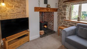 A cozy living room with a stone wall, a fireplace, a television on a wooden stand, and a grey sofa. - Wayside House (Craven Arms)