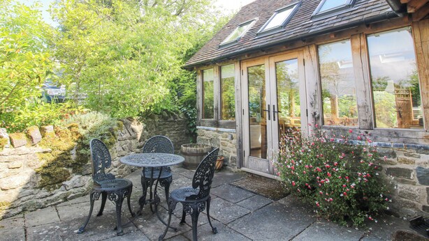 A stone building with a wooden exterior, a glass door, and a small patio with metal chairs and a table. - Wayside House (Craven Arms)