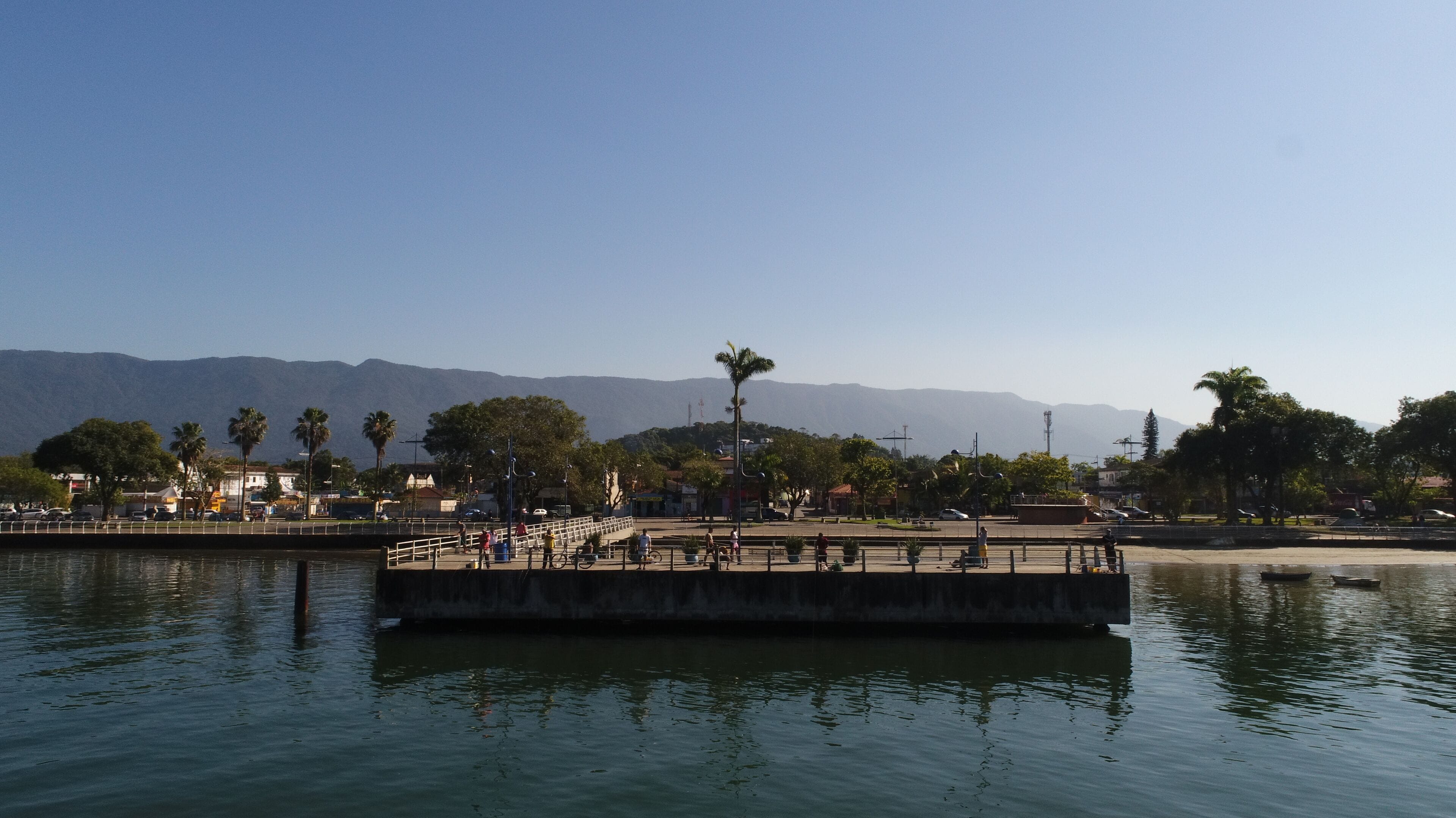 A waterfront promenade with a concrete pier, palm trees, and a mountain backdrop.