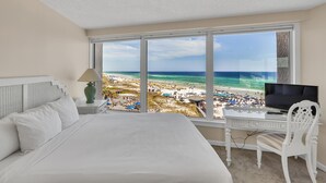 Ein Hotelzimmer mit großem Fenster mit Blick auf einen Strand, einem Bett, einem Schreibtisch mit Fernseher und einer Lampe.