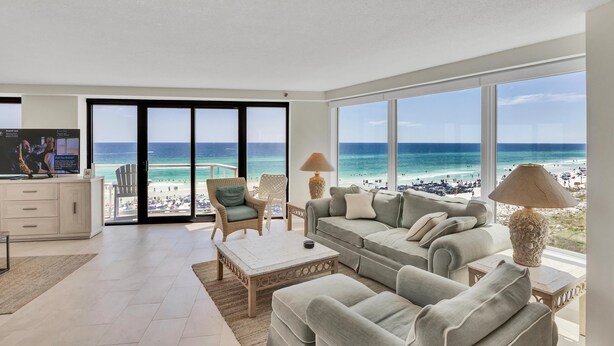 Ein großzügiger Wohnraum mit großem Fenster mit Blick auf einen Strand, ausgestattet mit einer Couch, Sesseln und einem Couchtisch.