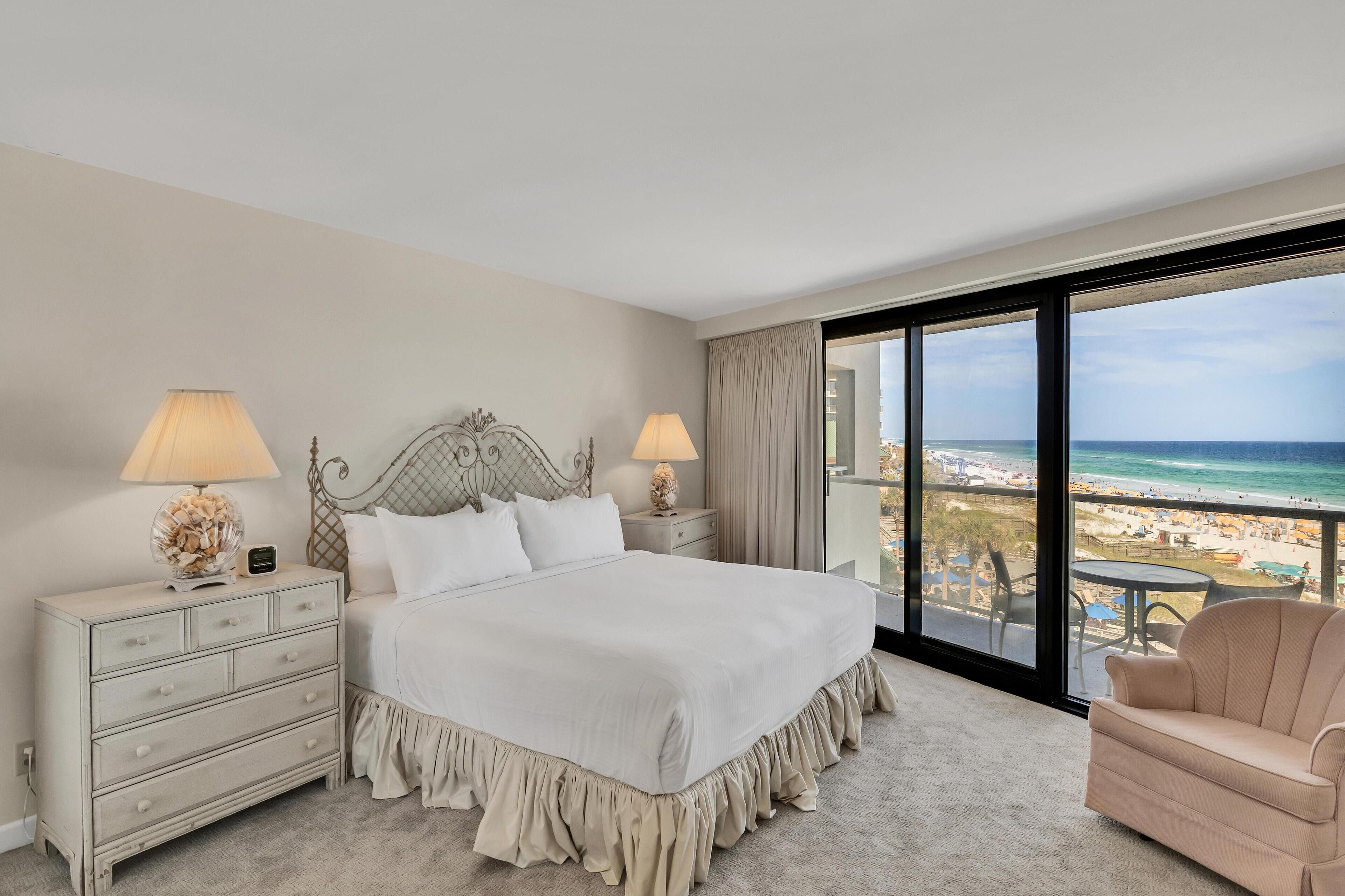 A hotel room with a large bed, two bedside lamps, a dresser, a chair, and a balcony with a view of the beach.