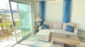 Un salon comprenant un canapé, un pouf et une table basse, une lampe, et une vue à travers une porte vitrée.