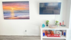Une pièce avec un grand tableau représentant un coucher de soleil sur l’océan, un téléviseur à écran plat et une console blanche agrémentée d’objets décoratifs et d’une inscription « FAMILLE ».