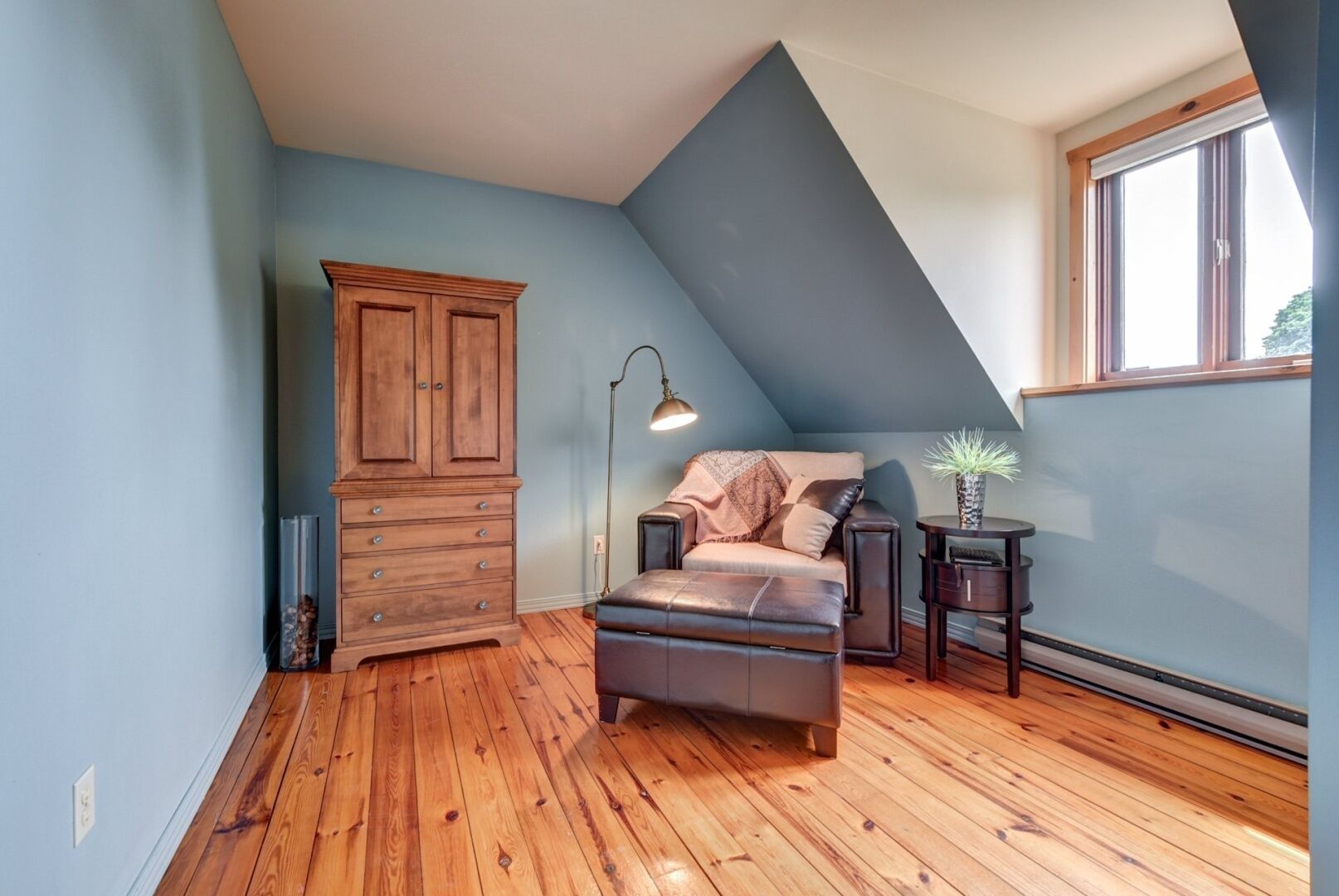 A loft bedroom with a sloped ceiling, wooden flooring, a large wardrobe, a sofa, a floor lamp, and a small table with a plant.