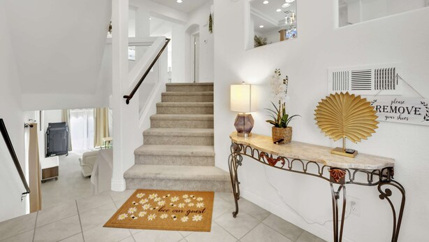 Uma escadaria com carpete bege, um tapete de entrada com um desenho de abelha, um abajur, um objeto decorativo na parede e uma placa indicando para retirar os sapatos.