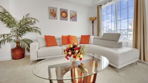 Uma sala de estar moderna com um sofá branco, almofadas laranja, uma mesa de centro de vidro e uma janela grande com cortinas.