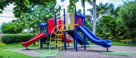 A playground with slides and climbing structures, surrounded by grass and trees.