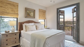 A bedroom with a bed, bedside tables, a lamp, a painting, and a large window with a view. - Sotol by Avantstay Direct River Access + Views (Dripping Springs)