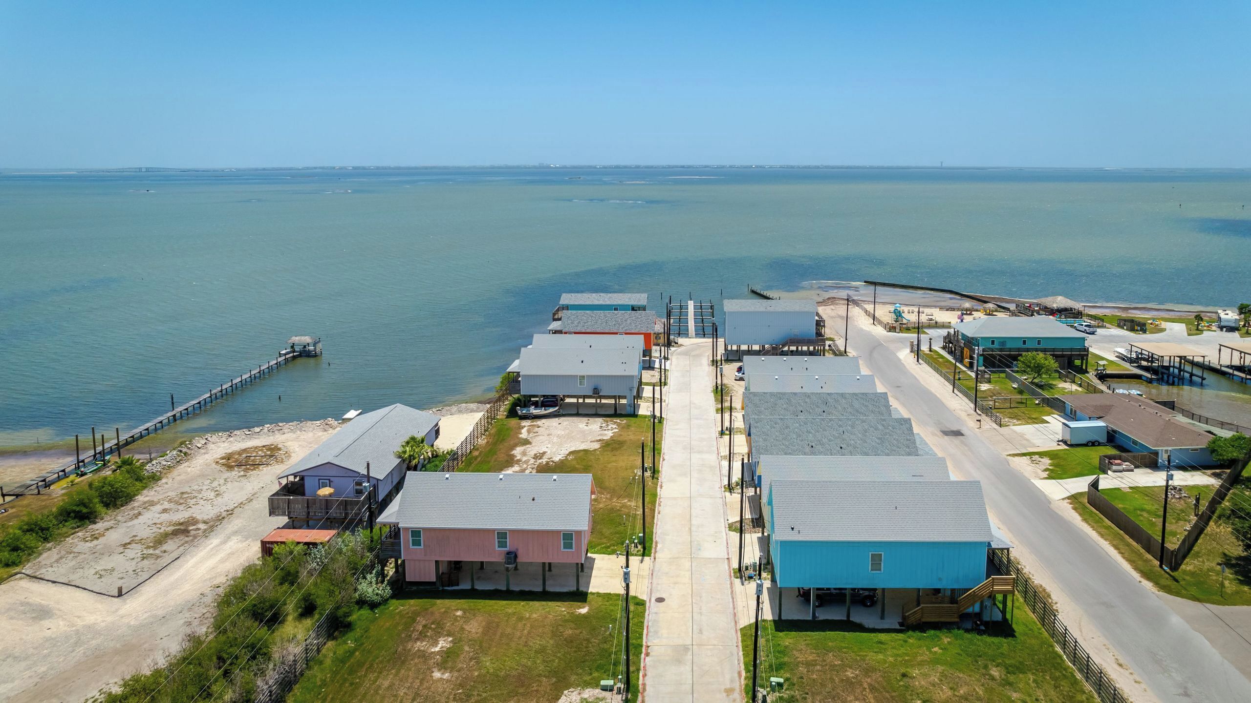 An aerial view of a coastal residential area with houses, a pier, and a clear view of the ocean.