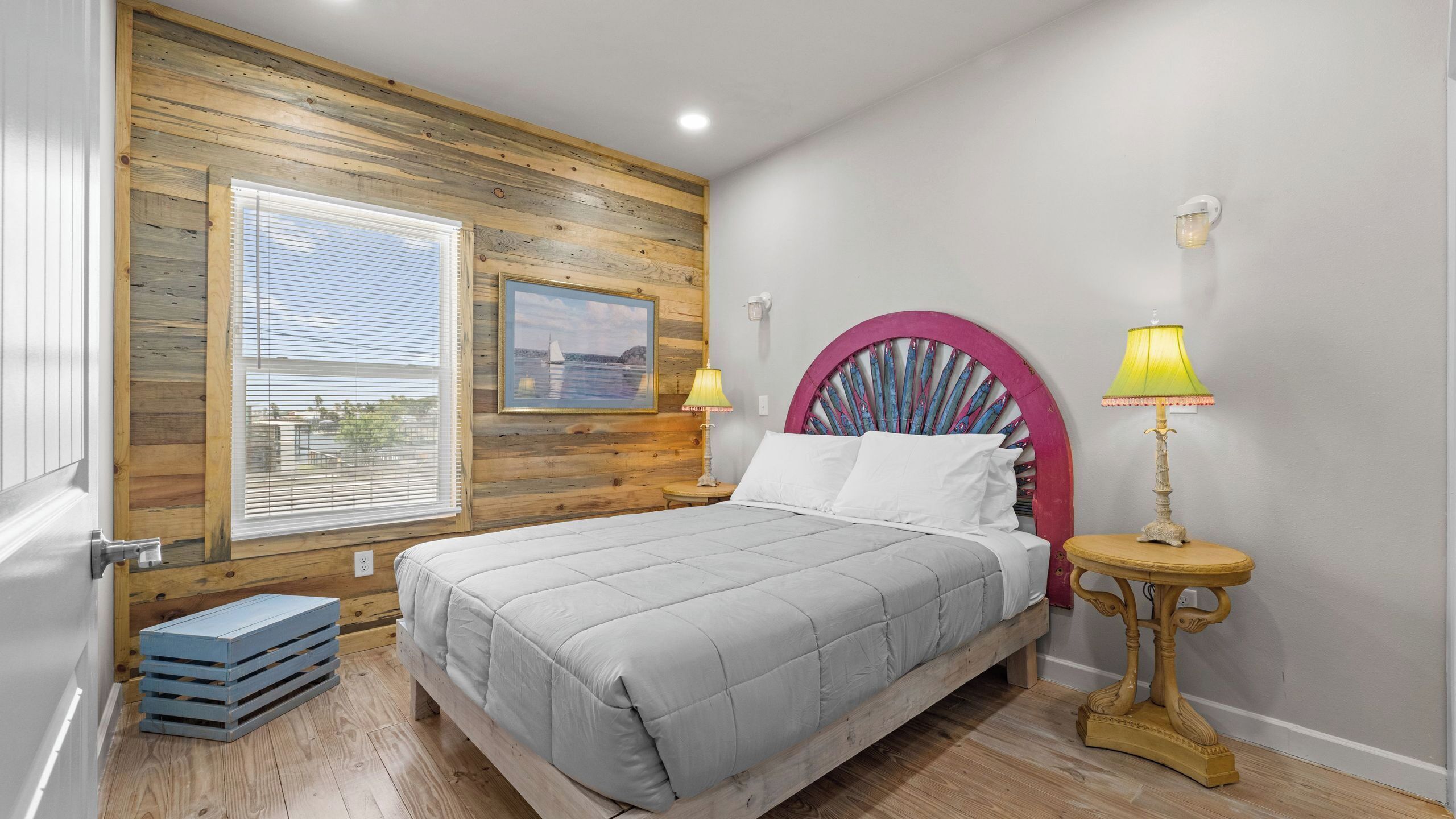 A bedroom with a wooden wall, a bed, a window with blinds, a painting, a lamp, and a small table.