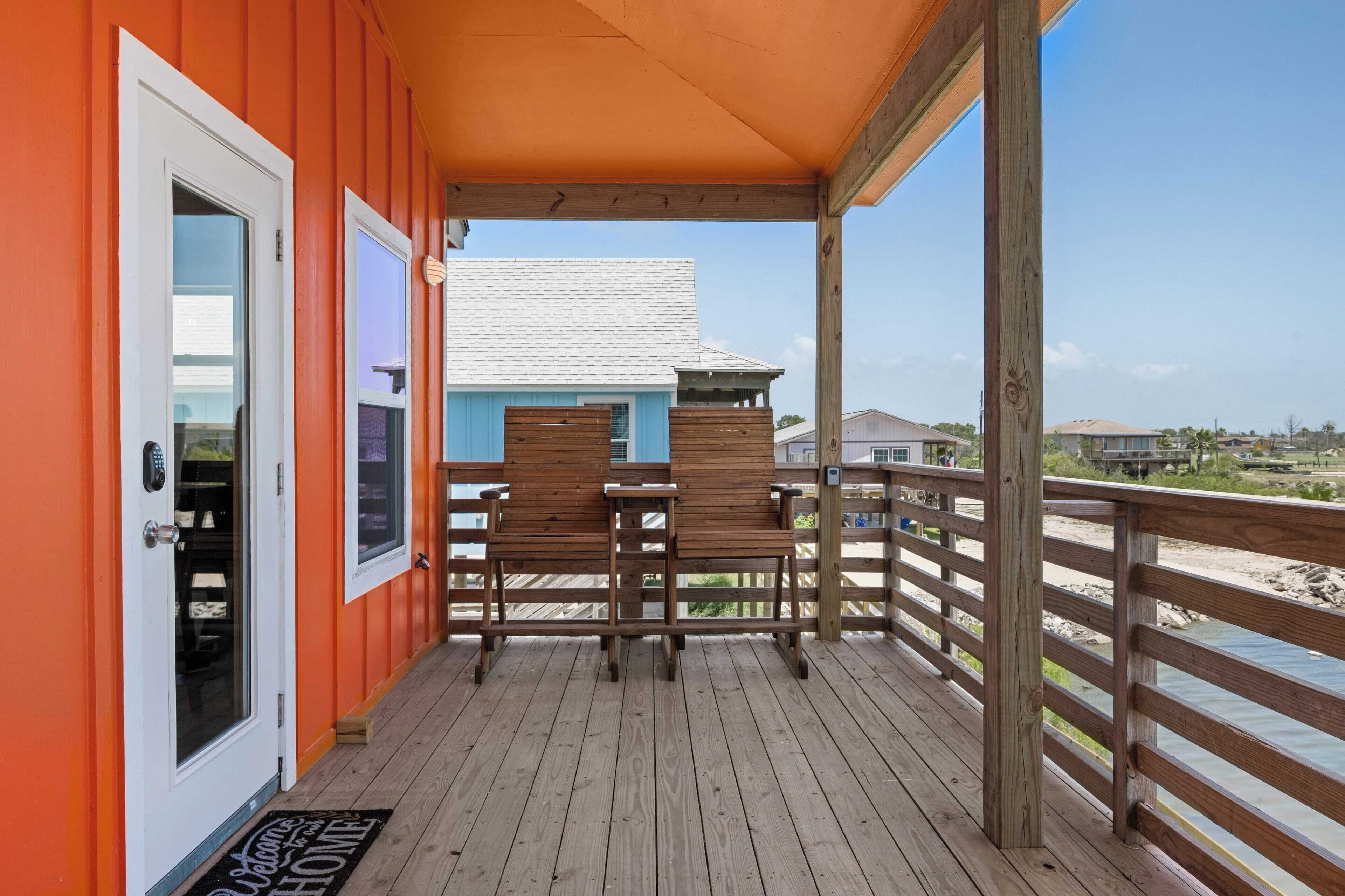 A wooden deck with two wooden benches, a door with a glass panel, and a view of a beach and buildings in the background.