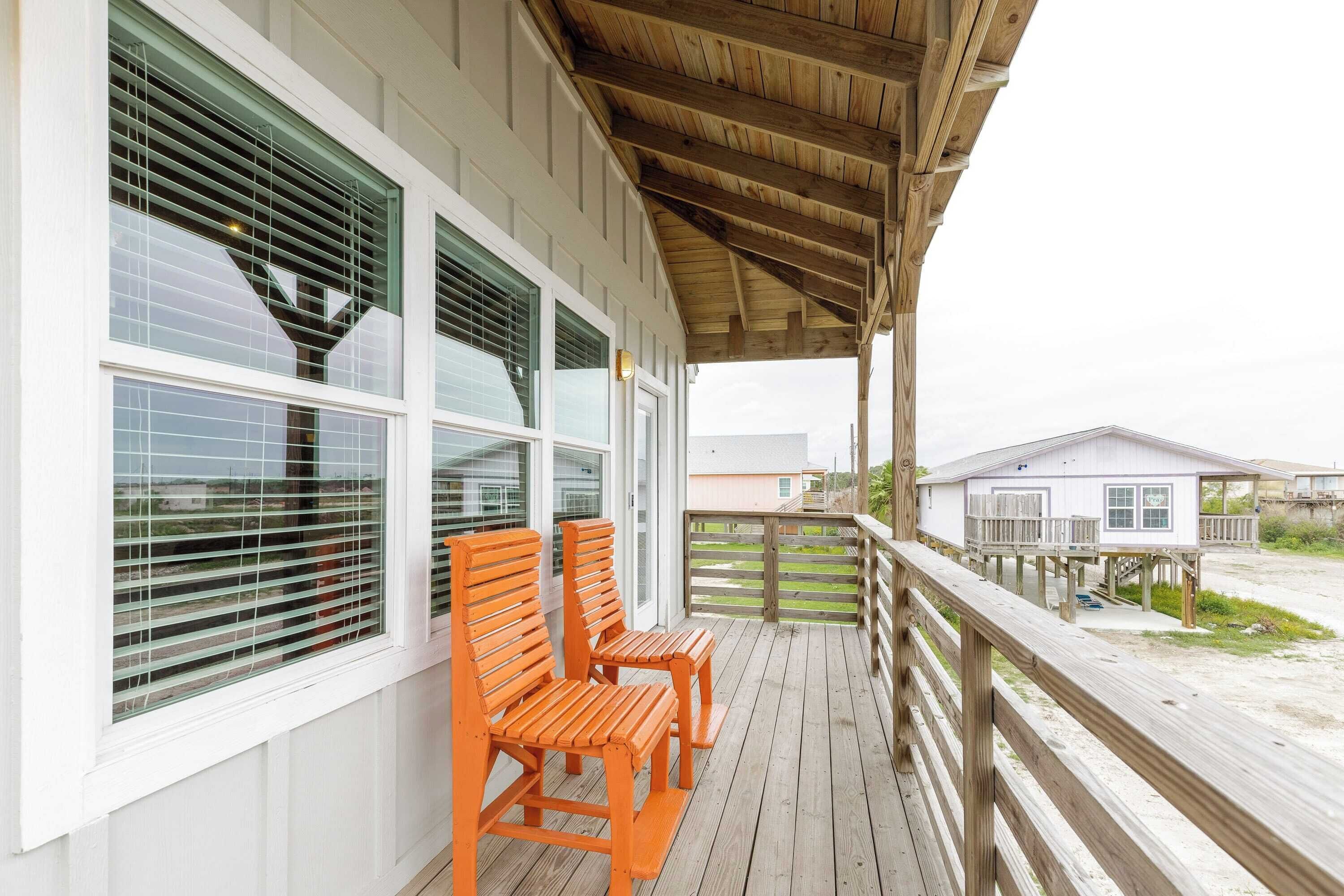 A wooden deck with two orange chairs, a covered area with a wooden railing, and a view of a beach house in the background.