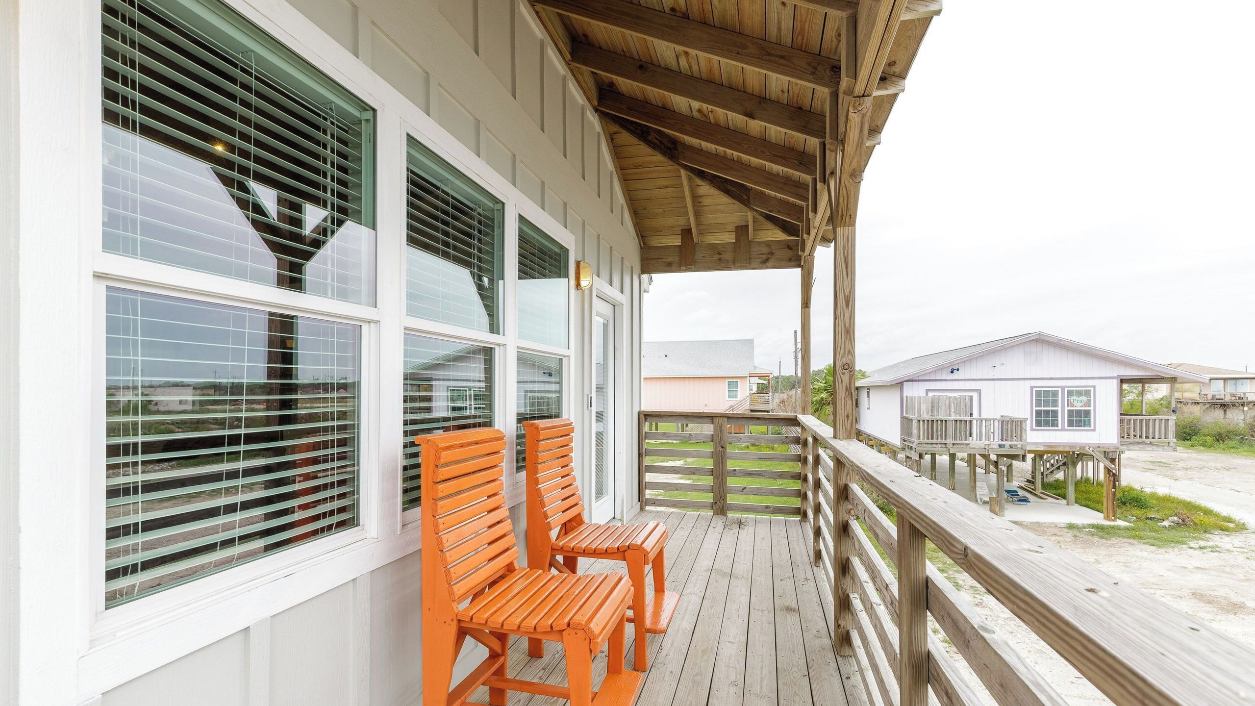 A wooden deck with a covered porch, large windows with blinds, and a view of other buildings.