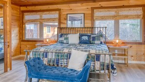Une chambre dans une cabane en bois, avec un grand lit, deux tables de chevet, un fauteuil bleu recouvert de tissu et de grandes fenêtres équipées de stores.