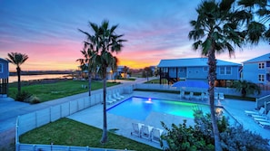 A pool area with a view of the sunset, palm trees, and a building with a blue roof. - Nature Preserve View, Shared Pool, Outdoor Living Casa Pescadora by Avantstay (Port Aransas)