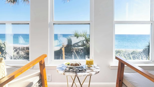 A room with a view of the ocean through large windows, a small table with breakfast items, and a wooden chair.