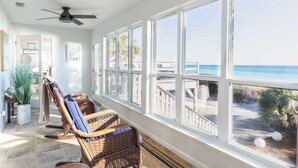 A sunlit room with large windows overlooking a beach, wicker chairs, a ceiling fan, and a potted plant.