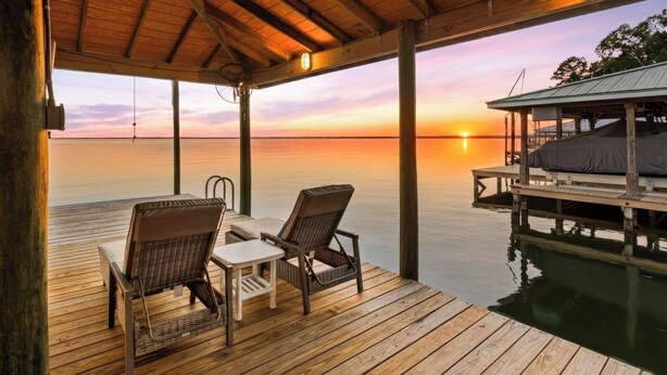 Een houten terras met rieten stoelen en een kleine tafel, met uitzicht op een rustig wateroppervlak tijdens de zonsondergang.
