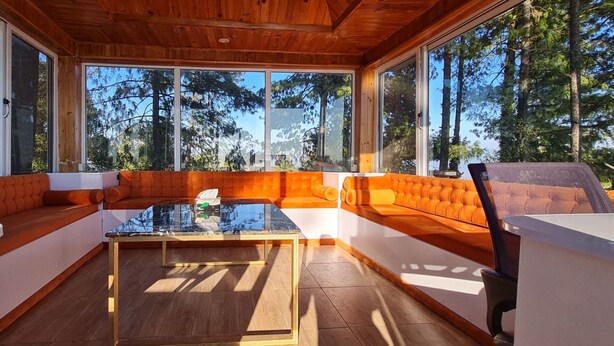 Interior moderno de cabaña con techo de madera, ventanas amplias, una mesa de centro de vidrio y asientos tapizados en color naranja.