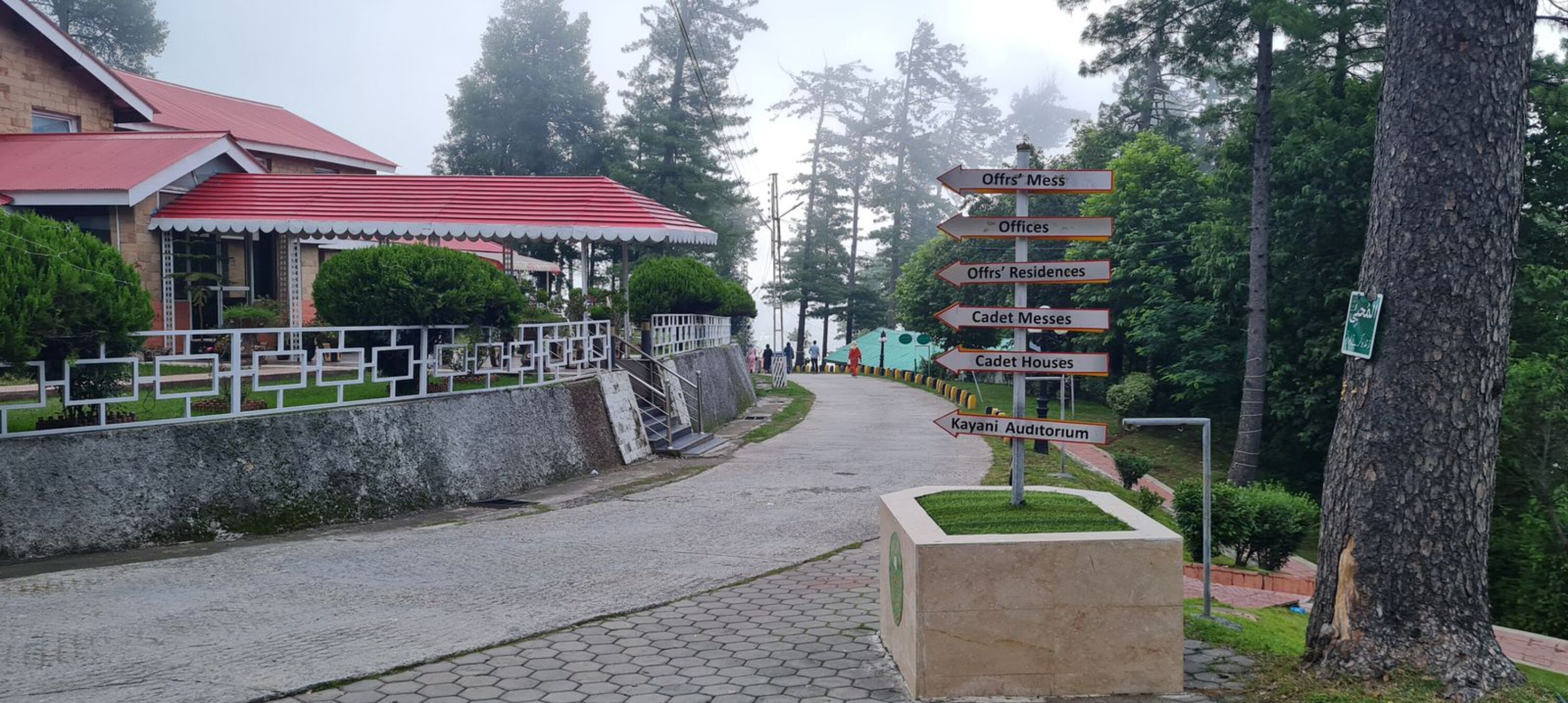 Un sendero conduce a un edificio con tejado rojo y un cartel indicador que muestra direcciones a varias instalaciones, incluyendo 'Offrs' Mess', 'Offrs' Residences', 'Cadet Messes', 'Cadet Houses' y 'Kayani Auditorium'.