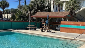 A clear blue pool with a 3 FT depth marker, surrounded by a sandy area and a wooden deck with picnic tables and umbrellas.