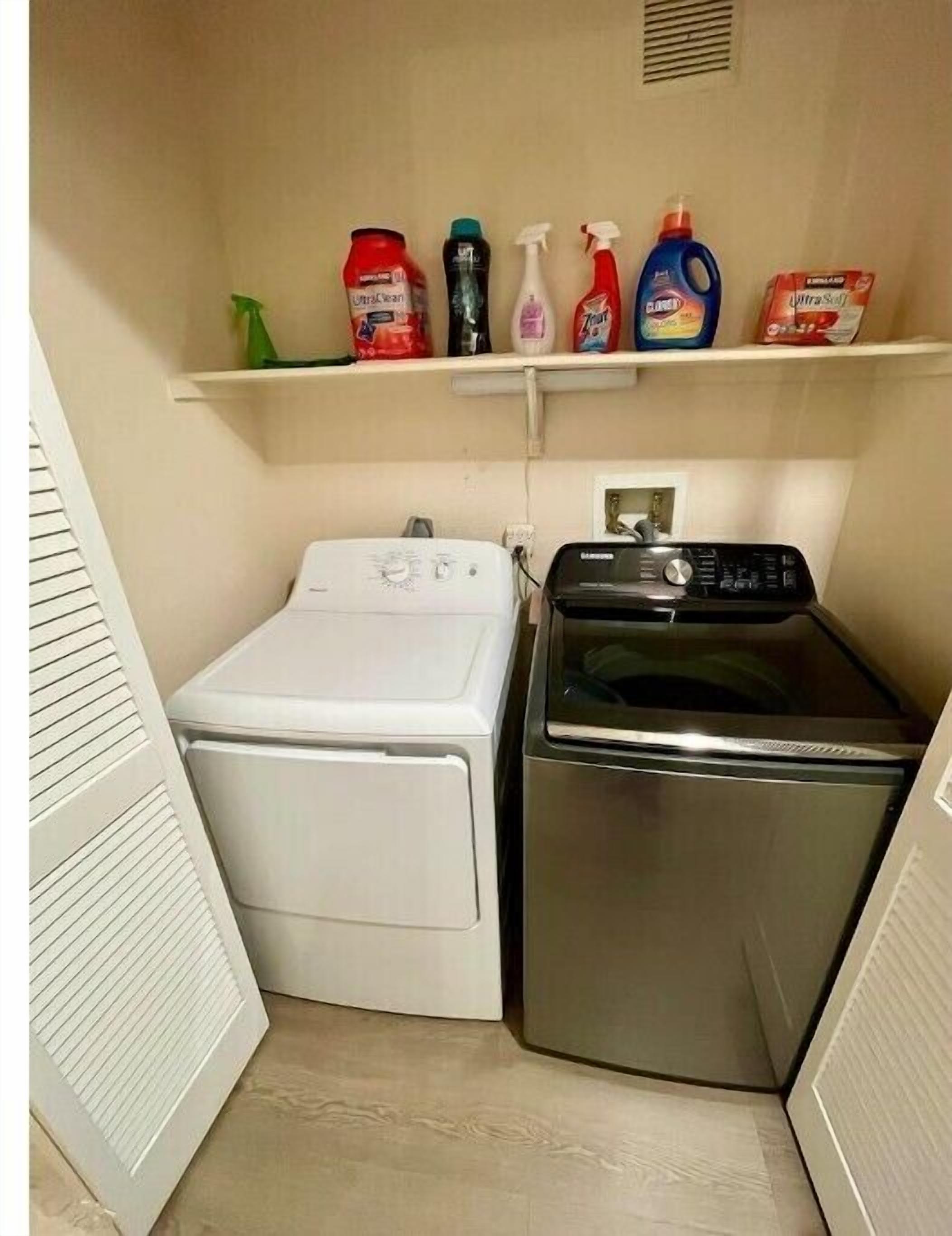 A compact laundry room with a white washer and dryer, a shelf with various cleaning products, and a vent above the dryer.