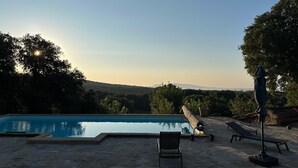 Una piscina con sillones de descanso y una pérgola, rodeada de árboles y un cielo despejado.