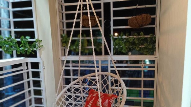 Una silla colgante de macramé blanco con un cojín rojo en un balcón estrecho con barandillas de metal y plantas en macetas.