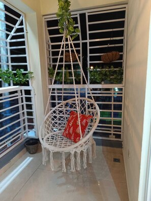 A white macrame hanging chair with a red cushion in a narrow balcony area with metal railings and potted plants.