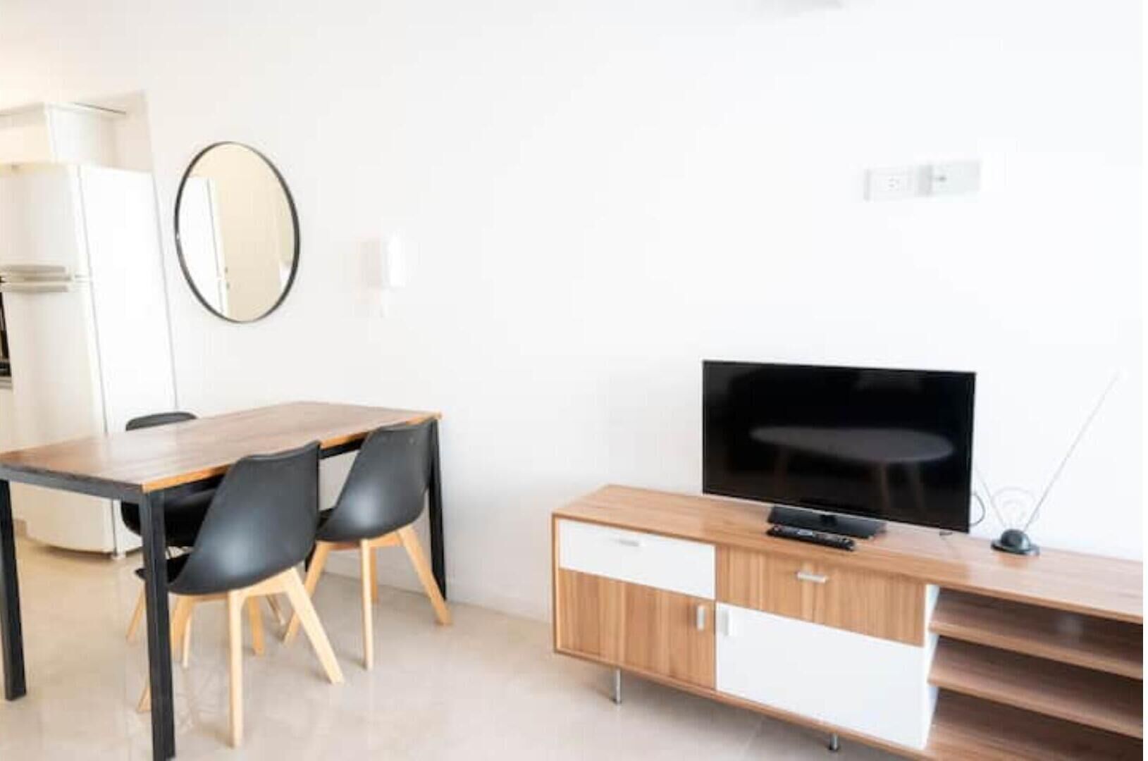 Un soggiorno moderno con un tavolo da pranzo in legno, due sedie nere, un televisore a schermo piatto su un supporto in legno e un mobile contenitore bianco con cassetti.