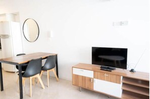 Um espaço de estar moderno com uma mesa de jantar de madeira, duas cadeiras pretas, uma televisão de tela plana em um suporte de madeira e um armário branco com gavetas.