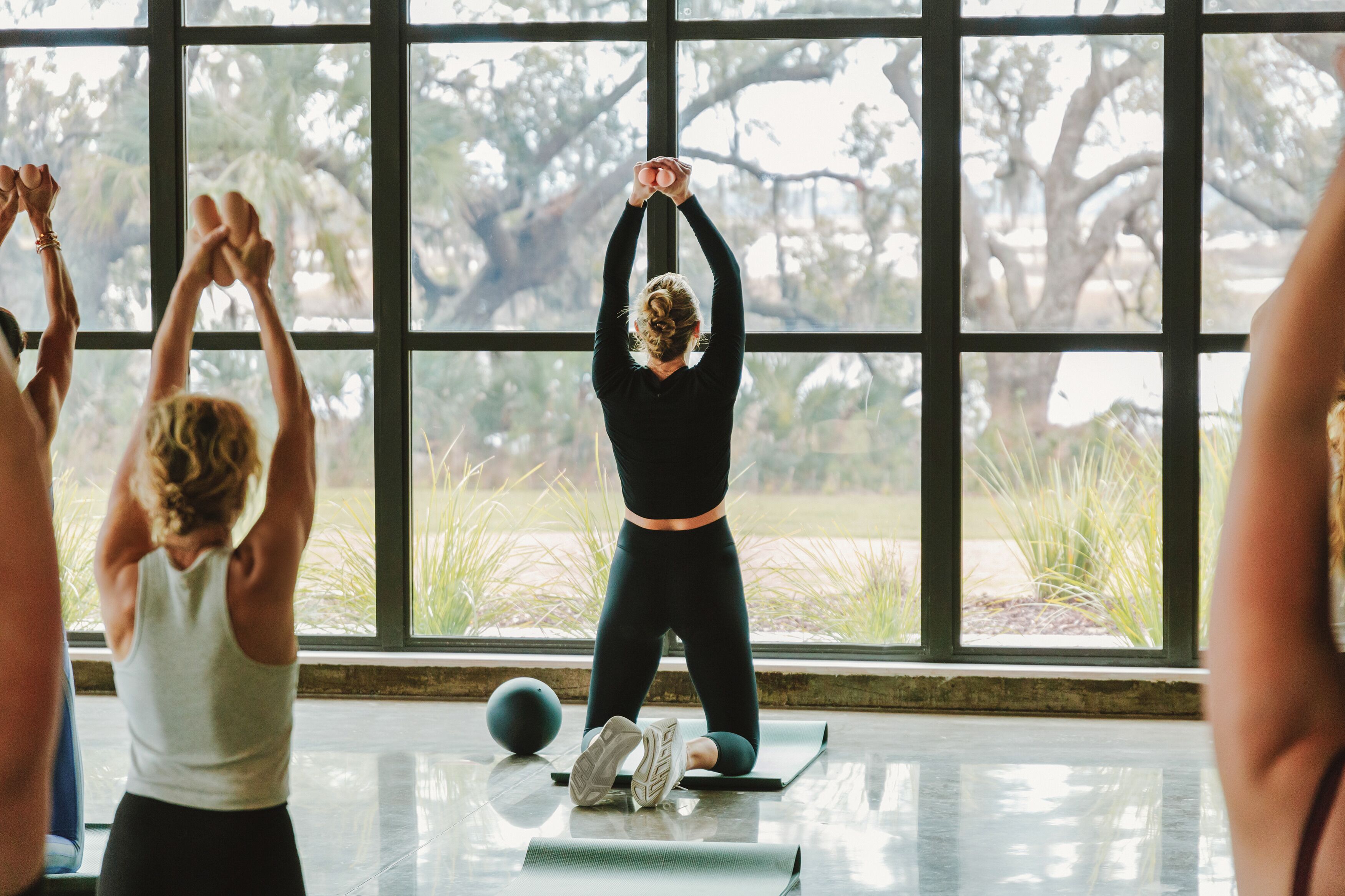 A group of people in a room with large windows, engaged in a yoga or stretching exercise.