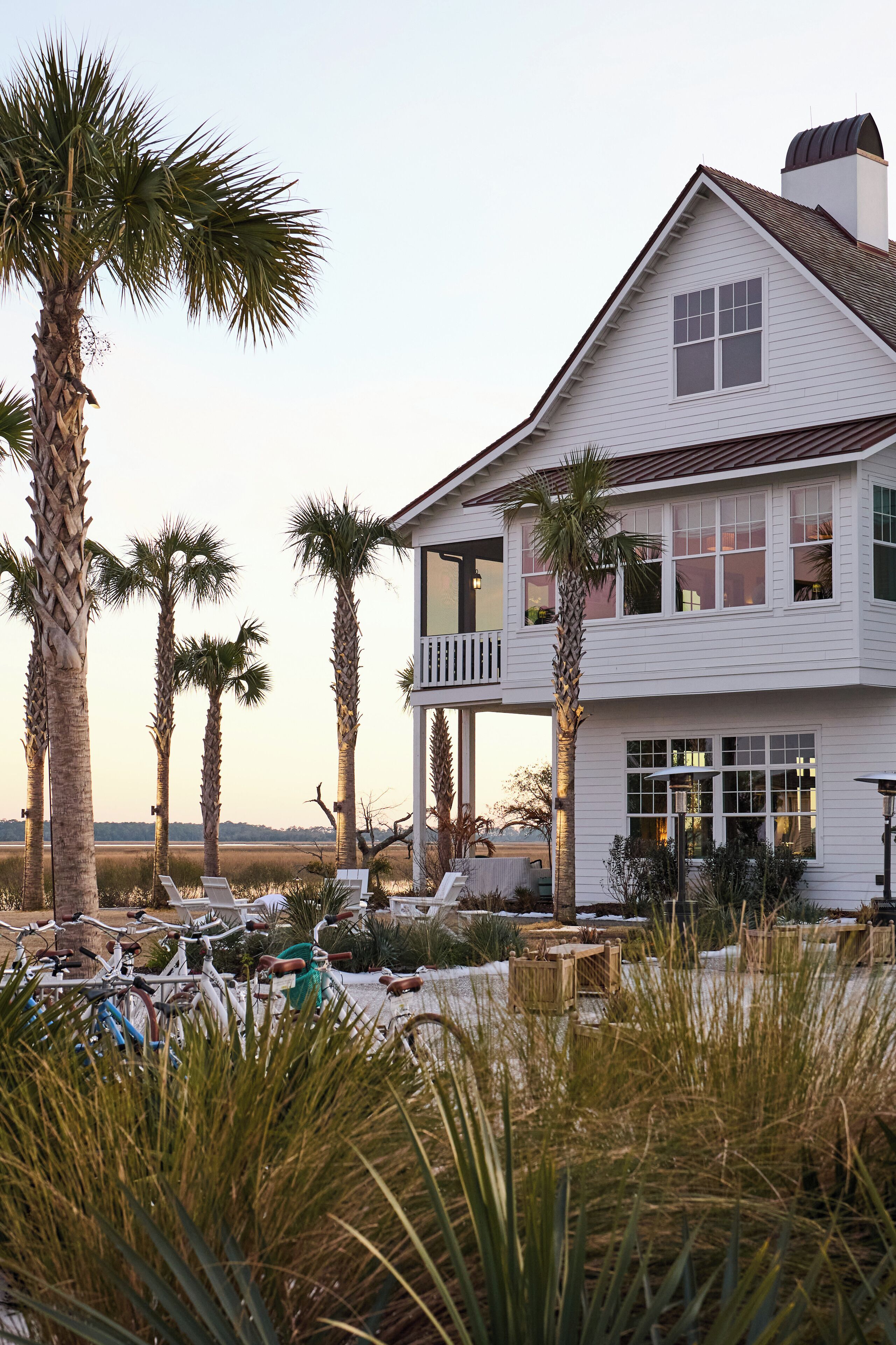A two-story white house with a balcony, surrounded by palm trees and a beach view.