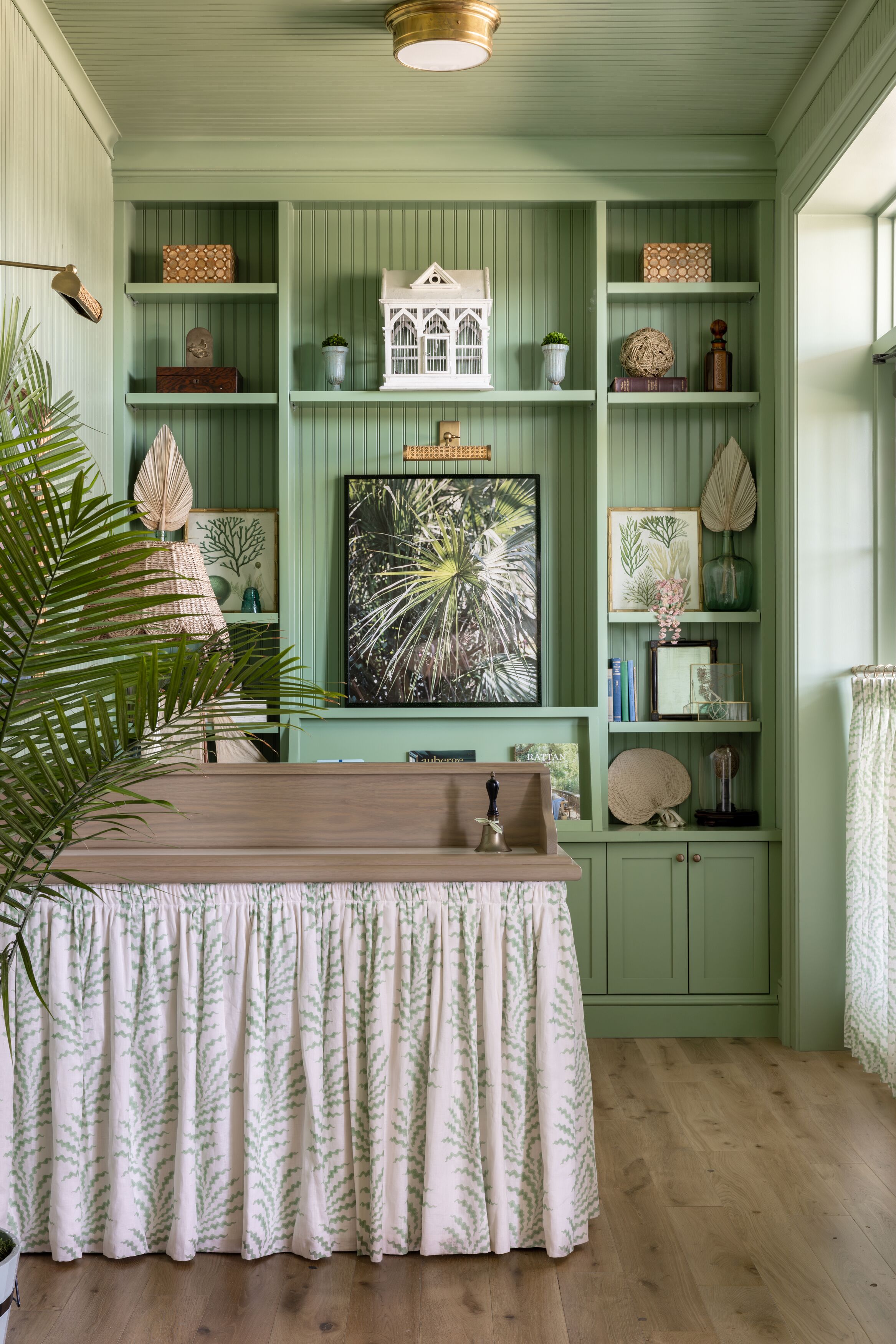 Een kamer met een groene wand, houten vloer en een tafel met een tafelkleed met een patroon. Er staan planken met diverse decoratieve items en een groot, ingelijst schilderij.
