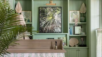 Een kamer met een groene wand, houten vloer en een tafel met een tafelkleed met een patroon. Er staan planken met diverse decoratieve items en een groot, ingelijst schilderij.